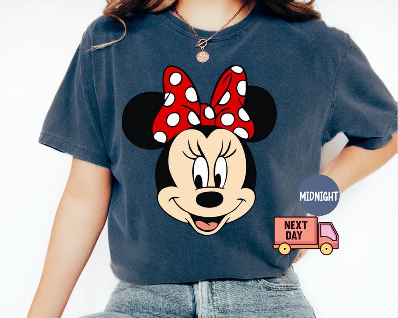 Może przedstawiać: Koszulka w ciemnoniebieskim kolorze z nadrukiem kresk&oacute;wkowej postaci Minnie Mouse. Minnie Mouse ma na sobie czerwoną kokardkę w białe kropki i szeroki uśmiech.