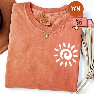 May include: Coral-colored t-shirt with a white sun graphic. The shirt has a crew neck and short sleeves. A gold necklace with a pendant is visible. The shirt is made of a soft material. The tag reads "Comfort Colors".