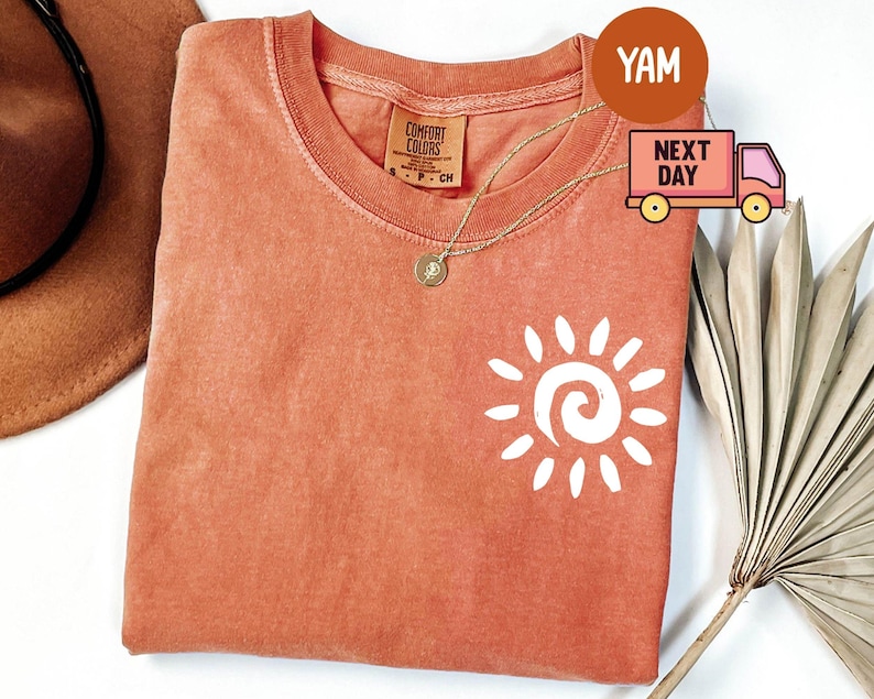 May include: Coral-colored t-shirt with a white sun graphic. The shirt has a crew neck and short sleeves. A gold necklace with a pendant is visible. The shirt is made of a soft material. The tag reads "Comfort Colors".