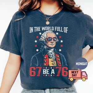 Op de afbeelding: Marineblauw T-shirt met een afbeelding van George Washington met een zonnebril met "1776" op de glazen. Het shirt heeft ook de tekst "IN THE WORLD FULL OF" en "67 BE A 76".