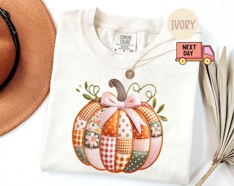 May include: Ivory-colored t-shirt featuring an embroidered pumpkin design. The pumpkin is composed of various fabric patterns in orange, pink, green, and brown, with a pink bow and green vines. The shirt has a Comfort Colors tag.
