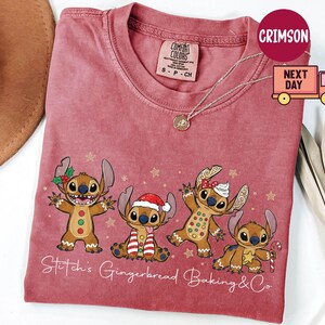 Puede incluir: Una camiseta roja Comfort Colors con un dise&ntilde;o festivo de cuatro personajes Stitch estilo pan de jengibre. La camiseta tiene el texto "Stitch's Gingerbread Baking & Co" en cursiva. Se ve un collar dorado.