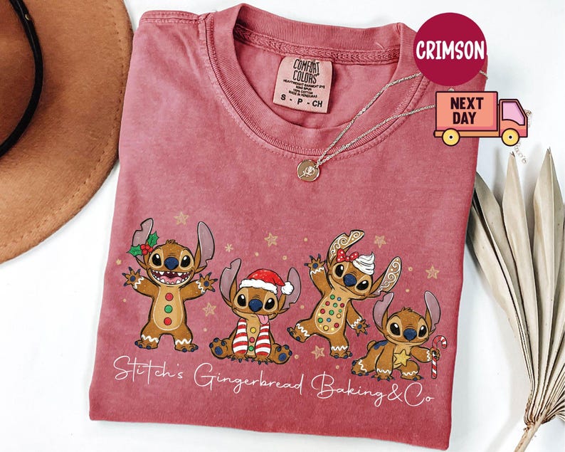 Puede incluir: Una camiseta roja Comfort Colors con un dise&ntilde;o festivo de cuatro personajes Stitch estilo pan de jengibre. La camiseta tiene el texto "Stitch's Gingerbread Baking & Co" en cursiva. Se ve un collar dorado.