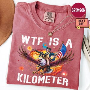May include: Crimson-colored t-shirt with the text "WTF IS A KILOMETER." The graphic depicts a person riding an eagle, holding a rifle and an American flag. The shirt has a crew neck and short sleeves.
