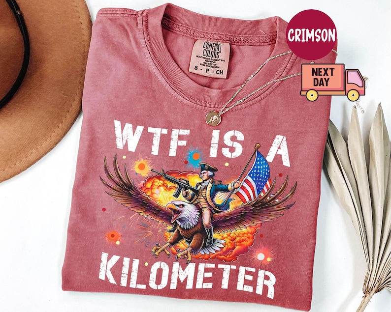 May include: Crimson-colored t-shirt with the text "WTF IS A KILOMETER." The graphic depicts a person riding an eagle, holding a rifle and an American flag. The shirt has a crew neck and short sleeves.