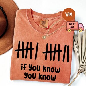 May include: Coral-colored t-shirt with black tally marks and the phrase "if you know you know" in a bold, sans-serif font. A gold necklace and a brown hat are visible. The shirt is made of Comfort Colors material.