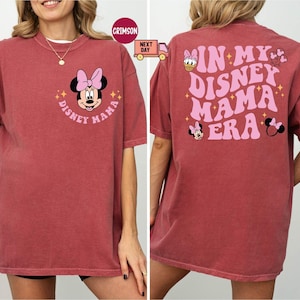 May include: A maroon t-shirt featuring Minnie Mouse with the text "Disney Mama" on the front. The back of the shirt displays the phrase "In My Disney Mama Era" with additional Disney character graphics.