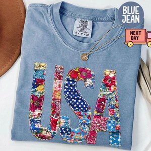May include: A light blue Comfort Colors t-shirt with the word "USA" appliqued on the front in a patchwork design of floral and star-spangled fabrics. A gold necklace with a pendant is draped on the shirt.