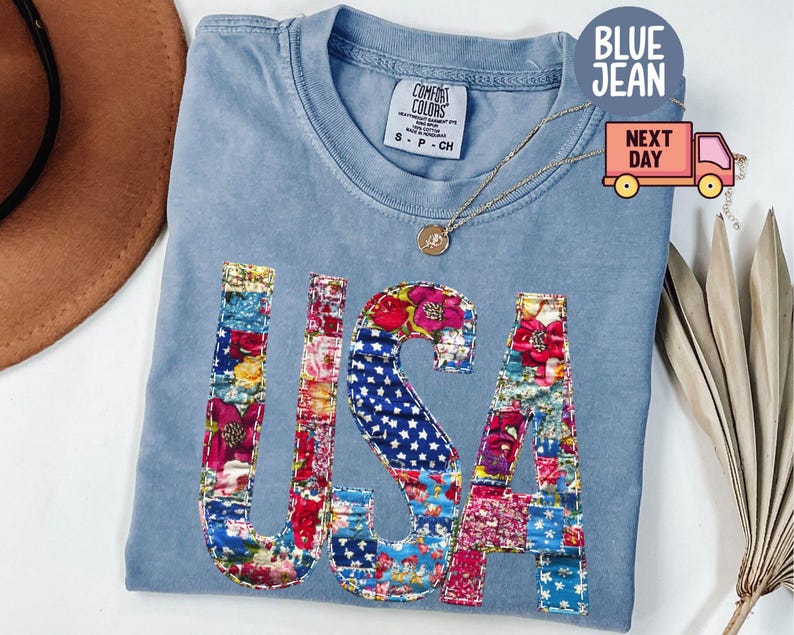 May include: A light blue Comfort Colors t-shirt with the word "USA" appliqued on the front in a patchwork design of floral and star-spangled fabrics. A gold necklace with a pendant is draped on the shirt.