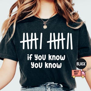 May include: Black t-shirt with white tally marks and the phrase "if you know, you know." The shirt has a crew neck and short sleeves. A black circle with the word "BLACK" is on the right side of the shirt.