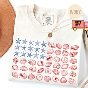 May include: An ivory short-sleeved t-shirt featuring a graphic print of a flag composed of blue starfish and red seashells. The shirt has a crew neckline and a Comfort Colors brand label at the inner collar. The design is arranged in a rectangular grid.