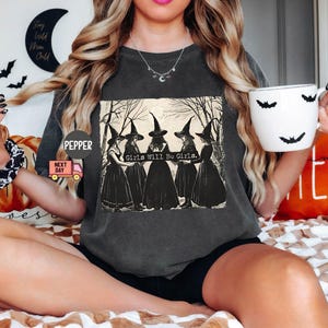 May include: A dark gray t-shirt with a graphic of four witches in black dresses and pointy hats. The text "Girls Will Be Girls." is printed below the witches.