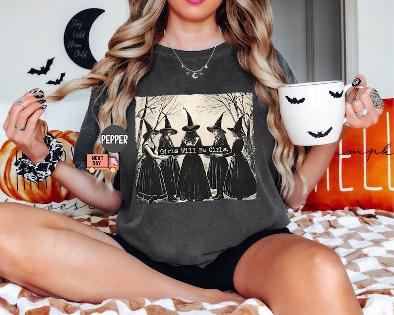 May include: A dark gray t-shirt with a graphic of four witches in black dresses and pointy hats. The text "Girls Will Be Girls." is printed below the witches.