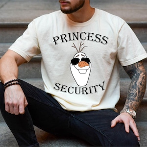 以下が含まれることがあります： サングラスとニンジンの鼻をした、アナと雪の女王のオラフのグラフィックが特徴のクリーム色のTシャツ。テキスト「PRINCESS SECURITY」は、オラフのグラフィックの上と下に黒で印刷されています。その人は黒いジーンズを履いています。