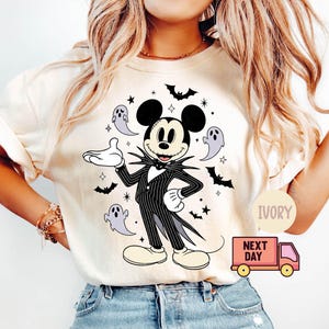 May include: Ivory-colored t-shirt featuring a graphic of Mickey Mouse dressed as Jack Skellington from The Nightmare Before Christmas. The design includes bats, stars, and ghosts. The shirt has short sleeves and a round neckline.