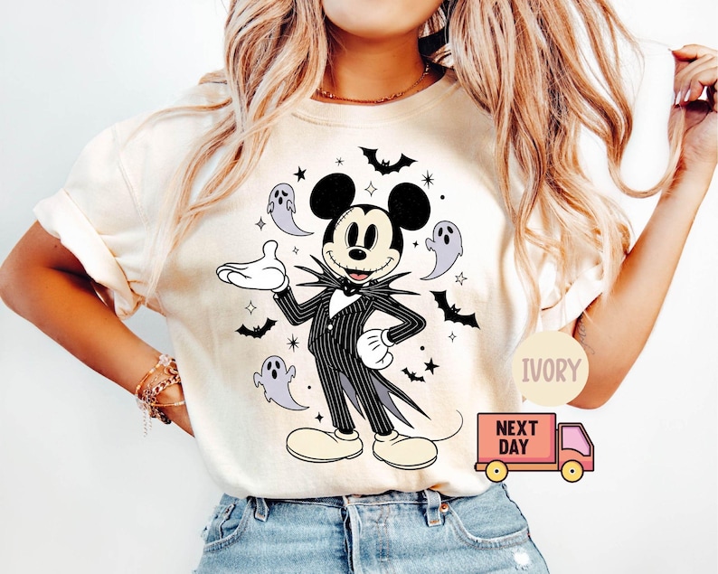 May include: Ivory-colored t-shirt featuring a graphic of Mickey Mouse dressed as Jack Skellington from The Nightmare Before Christmas. The design includes bats, stars, and ghosts. The shirt has short sleeves and a round neckline.