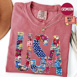 May include: A dusty rose Comfort Colors t-shirt with the word "USA" appliqued on the front. The letters are made of colorful floral and star-spangled fabric patches. A gold necklace is draped on the shirt.