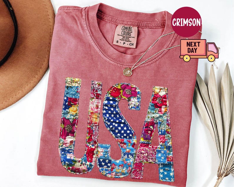 May include: A dusty rose Comfort Colors t-shirt with the word "USA" appliqued on the front. The letters are made of colorful floral and star-spangled fabric patches. A gold necklace is draped on the shirt.