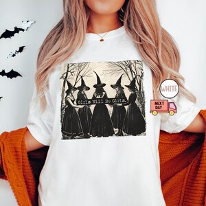 May include: White t-shirt with a black and white illustration of four witches in a forest. The witches are wearing black dresses and pointed hats. The text "Girls Will Be Girls." is printed on the shirt.