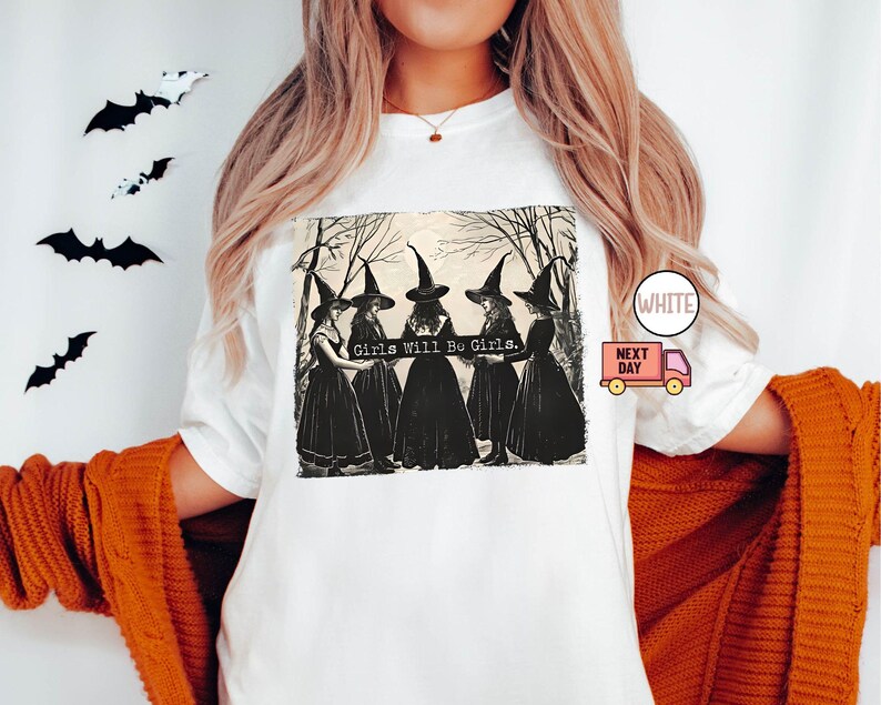 May include: White t-shirt with a black and white illustration of four witches in a forest. The witches are wearing black dresses and pointed hats. The text "Girls Will Be Girls." is printed on the shirt.