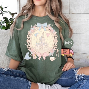K&ouml;nnte beinhalten: Ein gr&uuml;nes T-Shirt mit einem pastellrosa und blauen Motiv eines Schlosses, Ballons und einer Schleife. Das Motiv ist von einem floralen Design und einem wei&szlig;en Rahmen umgeben. Der Text "MOSS" und "NEXT DAY" ist auf dem Bild zu sehen.
