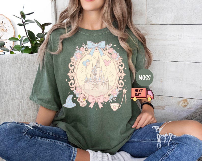 K&ouml;nnte beinhalten: Ein gr&uuml;nes T-Shirt mit einem pastellrosa und blauen Motiv eines Schlosses, Ballons und einer Schleife. Das Motiv ist von einem floralen Design und einem wei&szlig;en Rahmen umgeben. Der Text "MOSS" und "NEXT DAY" ist auf dem Bild zu sehen.