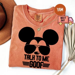 May include: A terracotta-colored t-shirt featuring a black graphic of a mouse silhouette wearing sunglasses. The text below the graphic reads TALK TO ME GOOF with horizontal lines. The shirt has a Comfort Colors label at the inner neckline.