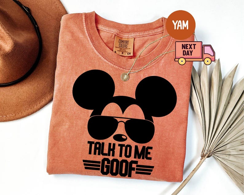 May include: A terracotta-colored t-shirt featuring a black graphic of a mouse silhouette wearing sunglasses. The text below the graphic reads TALK TO ME GOOF with horizontal lines. The shirt has a Comfort Colors label at the inner neckline.