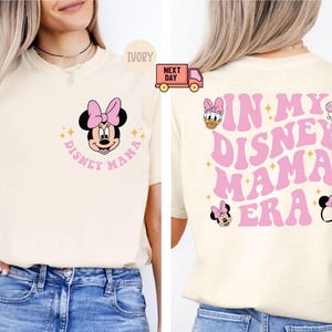 May include: Ivory-colored t-shirt featuring Minnie Mouse with a pink bow and the text "Disney Mama." The back of the shirt displays "In My Disney Mama Era" in pink, with additional Disney character graphics.