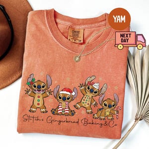 Puede incluir: Camiseta Comfort Colors de color coral con un gr&aacute;fico de cuatro personajes Stitch estilo pan de jengibre con decoraciones navide&ntilde;as. El texto "Stitch's Gingerbread Baking & Co" est&aacute; impreso debajo. Se ve un collar dorado.
