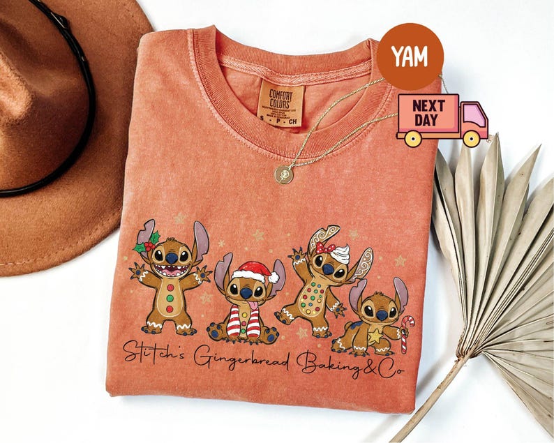 Puede incluir: Camiseta Comfort Colors de color coral con un gr&aacute;fico de cuatro personajes Stitch estilo pan de jengibre con decoraciones navide&ntilde;as. El texto "Stitch's Gingerbread Baking & Co" est&aacute; impreso debajo. Se ve un collar dorado.