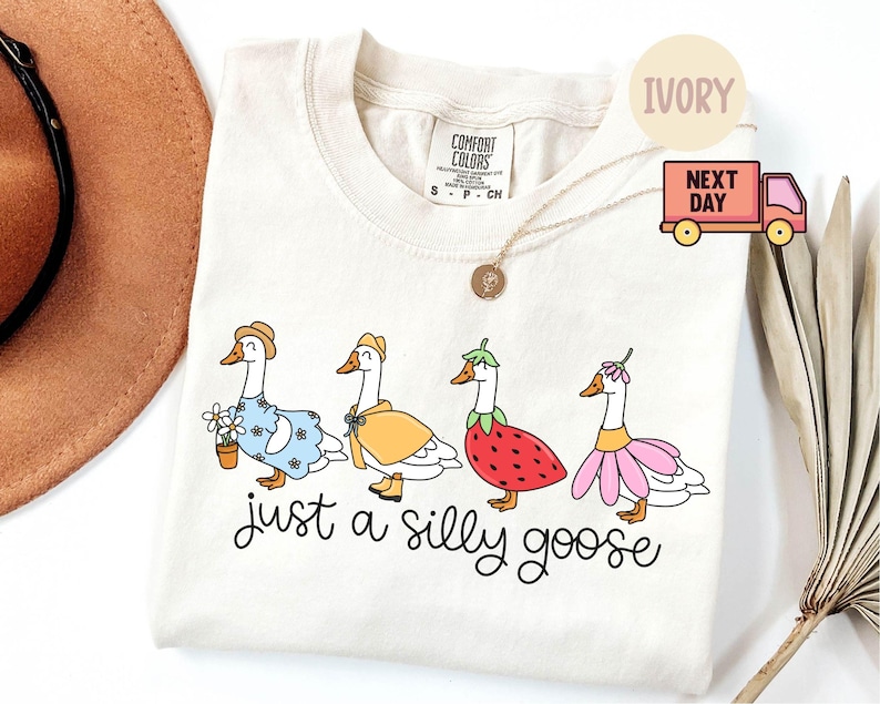 Peut inclure: T-shirt ivoire avec un motif fantaisiste de quatre oies de dessin anim&eacute; v&ecirc;tues de diff&eacute;rentes tenues. L'expression "just a silly goose" est &eacute;crite en script noir sous les oies. Le t-shirt a un col rond et des manches courtes.
