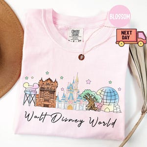 May include: A light pink t-shirt featuring a colorful illustration of Walt Disney World landmarks, including the Hollywood Tower Hotel and Cinderella's Castle. The words "Walt Disney World" are written in a script font below the graphic.