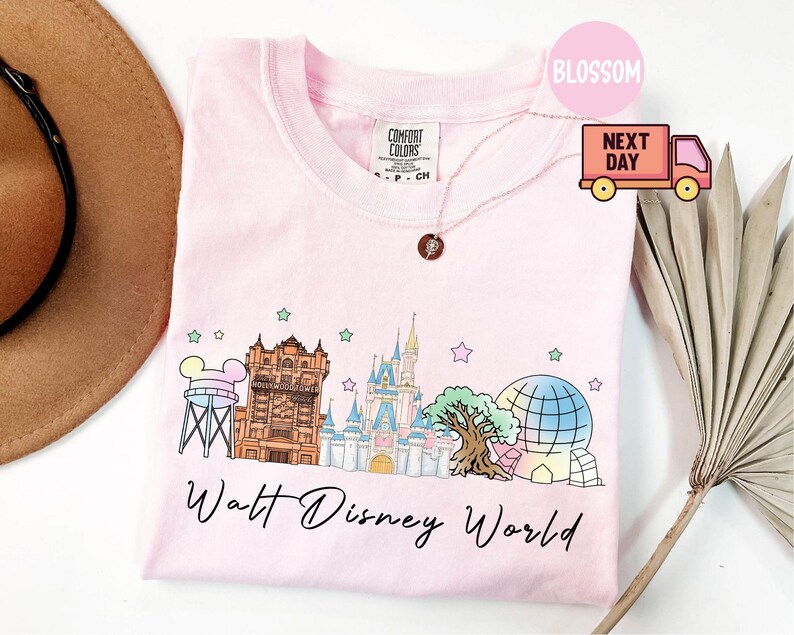May include: A light pink t-shirt featuring a colorful illustration of Walt Disney World landmarks, including the Hollywood Tower Hotel and Cinderella's Castle. The words "Walt Disney World" are written in a script font below the graphic.