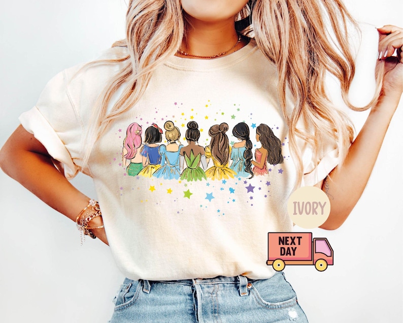 Puede incluir: Camiseta de color marfil con un gr&aacute;fico de siete princesas de dibujos animados de pie juntas. Las princesas llevan vestidos de colores y tienen diferentes colores de pelo. El gr&aacute;fico est&aacute; rodeado de estrellas.