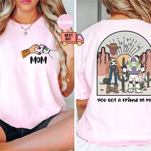Pu&ograve; includere: Una t-shirt rosa chiaro con una grafica frontale di due mani che formano un cuore con il testo MOM e una grafica sul retro di due personaggi dei cartoni animati in un paesaggio desertico con il testo YOU GOT A FRIEND IN ME stampato sotto.