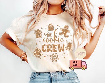 May include: Ivory-colored t-shirt with the text "The Cookie Crew" and festive cookie-themed graphics. The design includes a gingerbread man, house, gift, bell, Christmas tree, and snowflake. The shirt is paired with blue jeans.