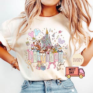 May include: Ivory-colored t-shirt featuring a colorful graphic of Disney characters, a castle, and the words "birthday girl." The design includes balloons, gifts, and a magic wand. The shirt has short sleeves.