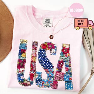 May include: A light pink t-shirt with the word "USA" in a patchwork design. The letters are filled with floral and star-spangled fabric. A gold necklace with a pendant is visible. The shirt is folded on a white surface.