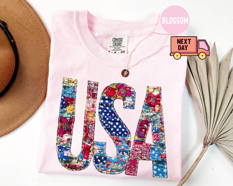 May include: A light pink t-shirt with the word "USA" in a patchwork design. The letters are filled with floral and star-spangled fabric. A gold necklace with a pendant is visible. The shirt is folded on a white surface.