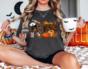 May include: A dark gray t-shirt with a graphic design of a stethoscope, a syringe, a pumpkin, and the word "NURSE" in orange and black letters. The graphic design is surrounded by fall leaves and pine cones.