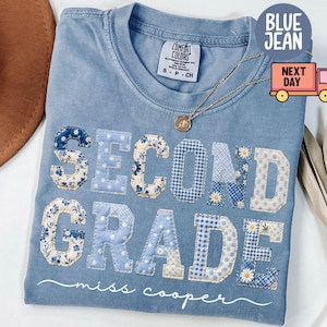 May include: A blue jean colored t-shirt featuring the text SECOND GRADE in large, fabric-appliqued letters with various blue and white floral, polka dot, and gingham patterns. Below the text, the name miss cooper is printed in white script.