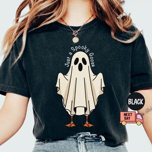 May include: Black t-shirt featuring a cartoon ghost with the text "Just a Spooky Goose". The ghost is white with black eyes and a beak, and orange feet. The shirt has a vintage look.