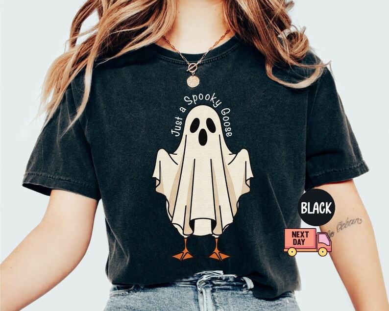 May include: Black t-shirt featuring a cartoon ghost with the text "Just a Spooky Goose". The ghost is white with black eyes and a beak, and orange feet. The shirt has a vintage look.