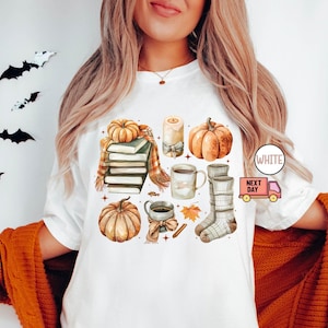 May include: White t-shirt with a fall-themed design featuring pumpkins, books, a mug of coffee, and a candle. The design is in a watercolour style.