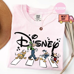 K&ouml;nnte beinhalten: Hellrosa T-Shirt mit dem Wort "Disney" in schwarzer Schrift und den Zeichentrickfiguren Pluto, Goofy, Donald Duck und Mickey Mouse, die einen Zebrastreifen &uuml;berqueren. Das Shirt hat einen Rundhalsausschnitt und kurze &Auml;rmel.