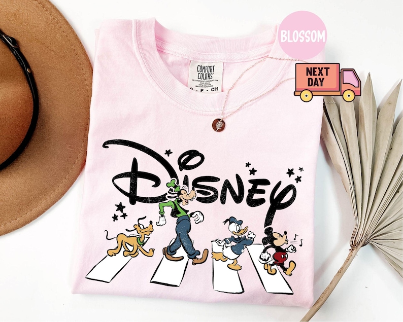 K&ouml;nnte beinhalten: Hellrosa T-Shirt mit dem Wort "Disney" in schwarzer Schrift und den Zeichentrickfiguren Pluto, Goofy, Donald Duck und Mickey Mouse, die einen Zebrastreifen &uuml;berqueren. Das Shirt hat einen Rundhalsausschnitt und kurze &Auml;rmel.