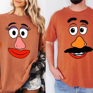 Könnte beinhalten: Zwei rostfarbene T-Shirts, jeweils mit einem Cartoon-Gesicht. Ein Shirt zeigt das Gesicht einer weiblichen Figur mit großen Augen, einer rosa Nase und roten Lippen. Das andere Shirt zeigt das Gesicht einer männlichen Figur mit schwarzem Schnurrbart und oranger Nase.