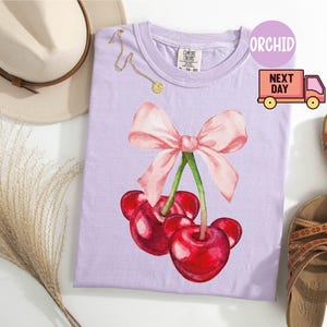 Puede incluir: Una camiseta de color lila claro con un dise&ntilde;o de acuarela de cerezas y un lazo rosa. La camiseta est&aacute; junto a un sombrero beige, un collar dorado y sandalias marrones. La palabra "ORCHID" est&aacute; impresa en la camiseta.