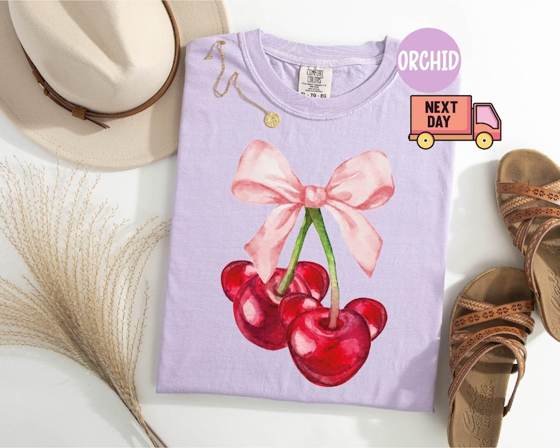 Puede incluir: Una camiseta de color lila claro con un dise&ntilde;o de acuarela de cerezas y un lazo rosa. La camiseta est&aacute; junto a un sombrero beige, un collar dorado y sandalias marrones. La palabra "ORCHID" est&aacute; impresa en la camiseta.